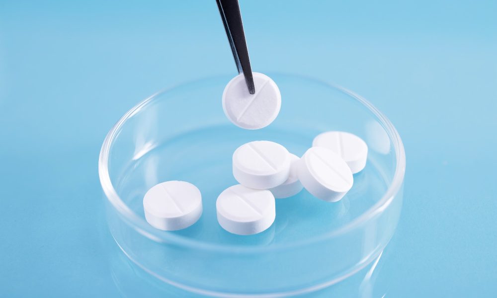 A closeup shot of tweezers picking up a white pill from a pile in a glass dish at a lab